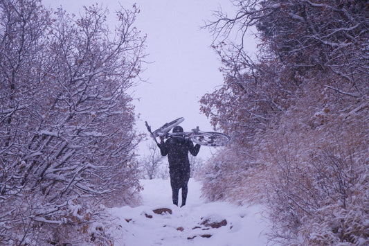 image of a man walking up a snowy trail in a blizzard with a bike over his shoulders and shrubs on either side of the trail.