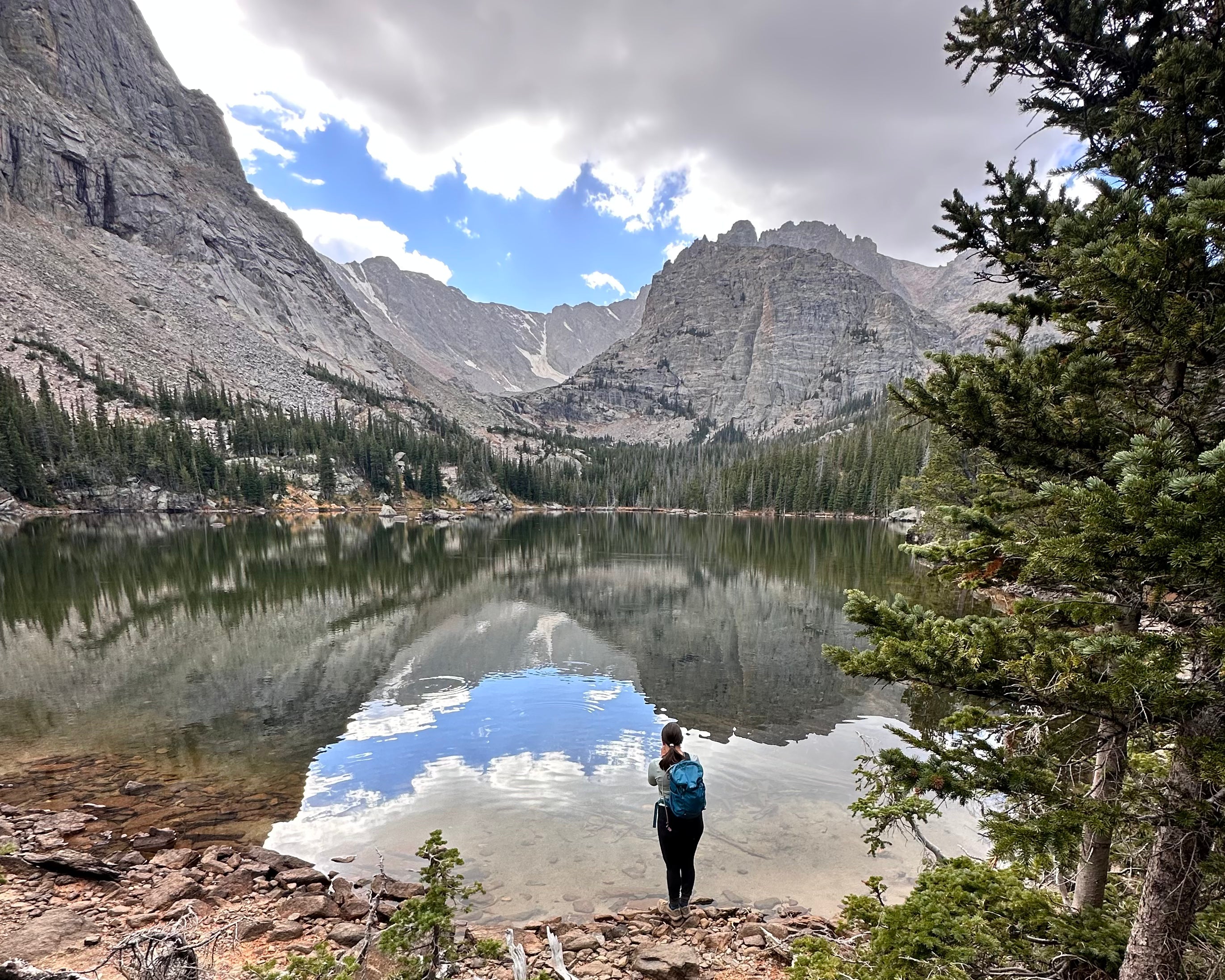 woman standing in frontt of a reflective late with small ripples across it. In the background there are pine trees crawling up the rocky hill which truns into a vast, wide, rocky mountain range. There is still evidence of snowy glaicers in some of the crags and cracks.